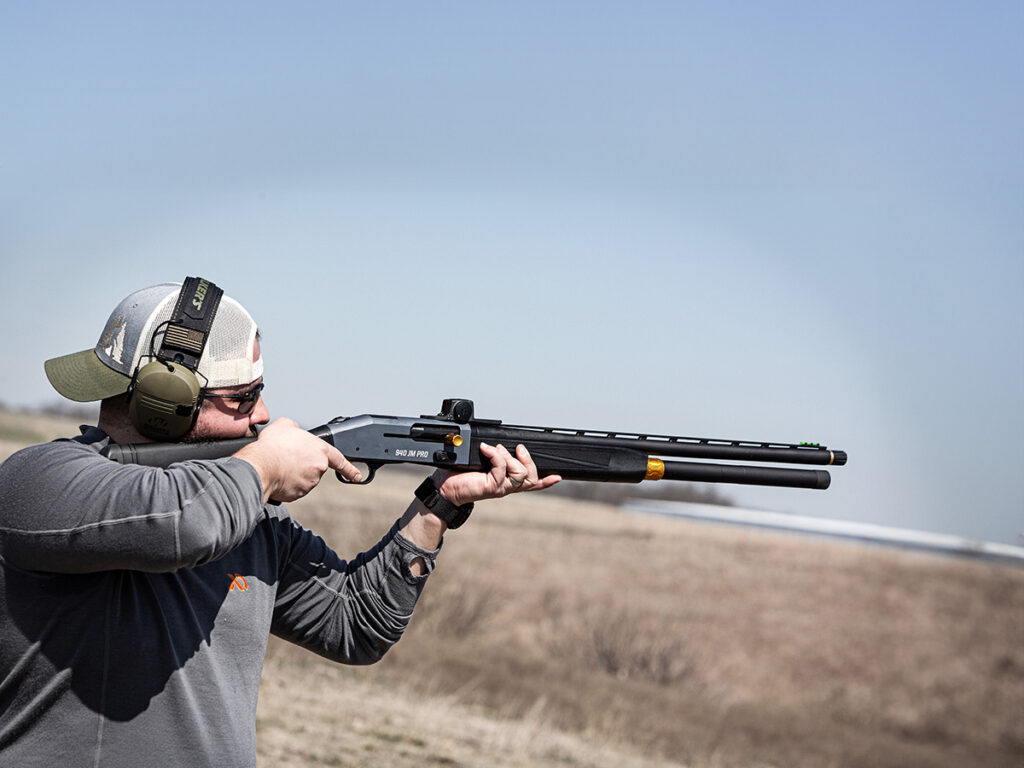A man shouldering a Mossberg 940 JM Pro shotgun in a field.