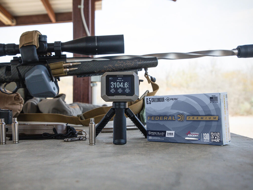 A box of Federal 6.5 Creedmoor +Peak ammo next to a rifle and a chronograph showing 3,104.6 fps.