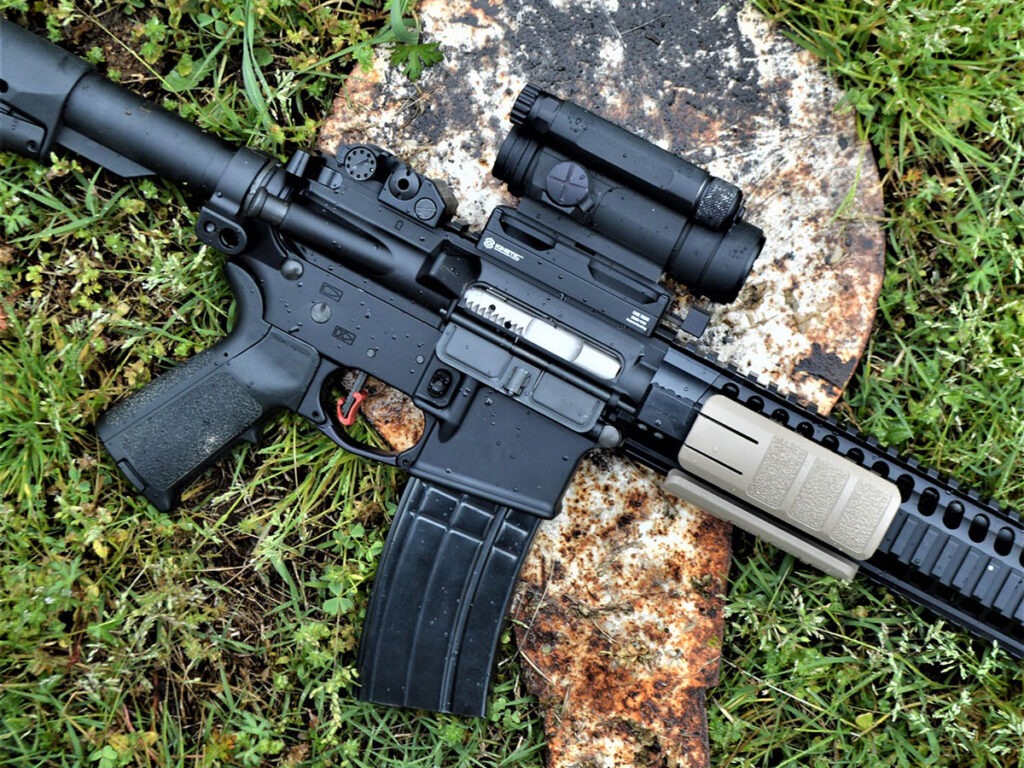 A modern AR-15 equipped with an Aimpoint red dot sight laying on a steel target in the grass.