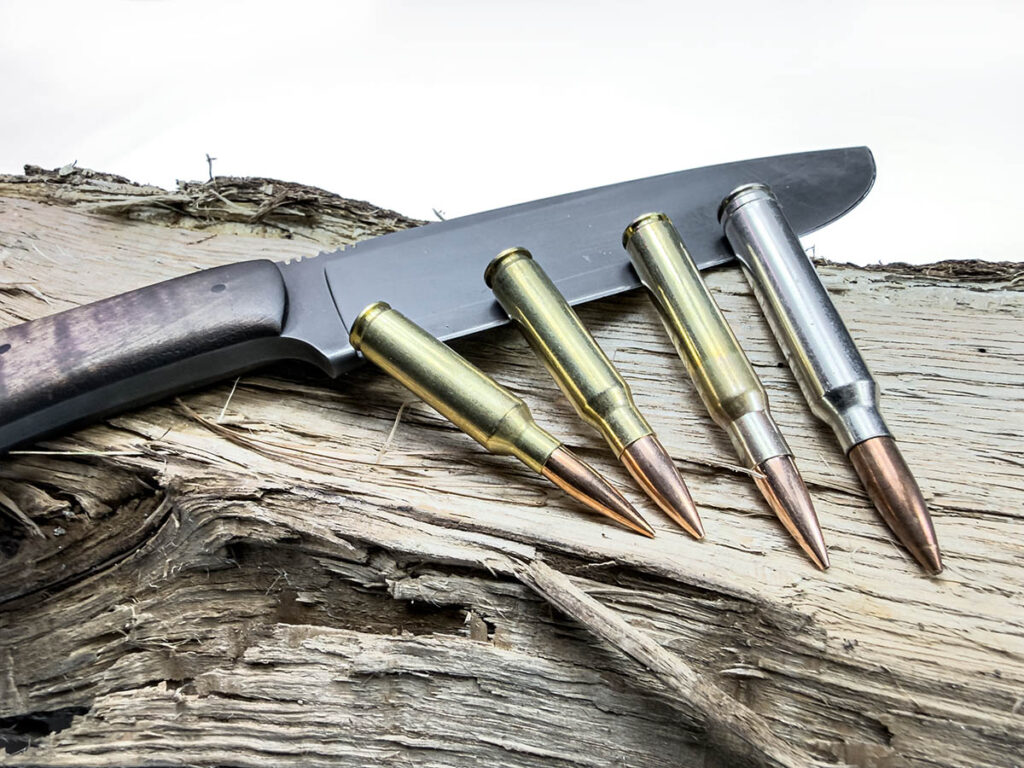 A 6.5 Creedmoor, .308 Win, .30-06, and .300 Win Mag cartridge lying on a knife on a log.