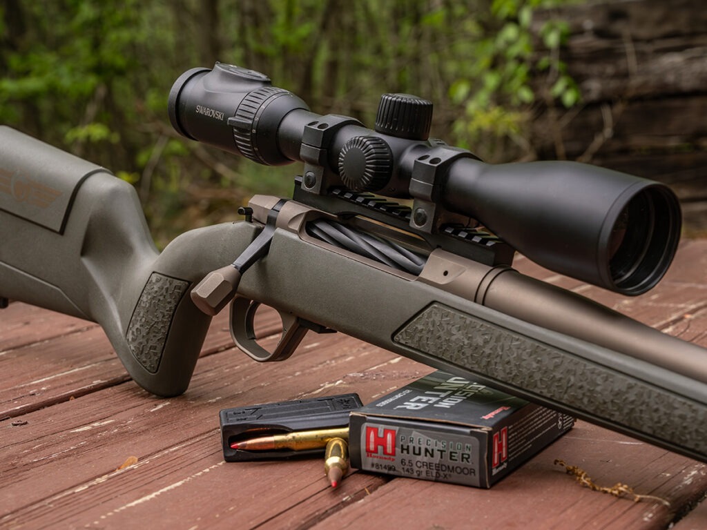 A scoped bolt-action rifle next to high bc 6.5 creedmoor bullets on a table.
