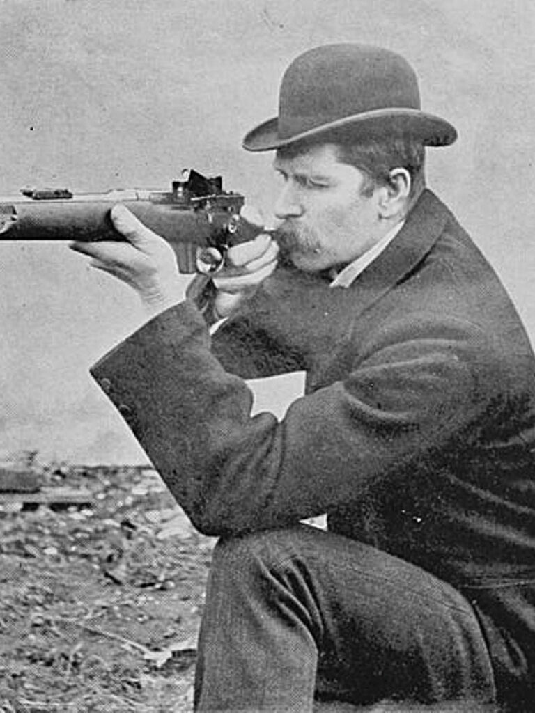 An old black and white photo of a man aiming a rifle equipped with a Grubb sight.