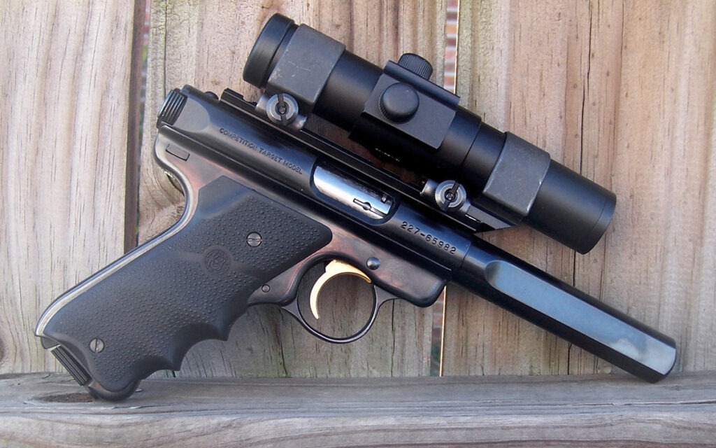 A Ruger .22 target pistol equipped with an early red dot sight leaning on a wooden fence.
